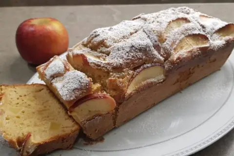 Un delizioso plumcake alle mele, cosparso di zucchero a velo, con una mela rossa sullo sfondo.