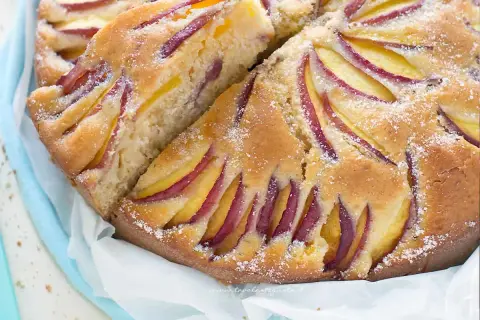 Una fetta di torta di pesche morbidissima, con pesche affettate disposte a raggiera sulla superficie dorata e cosparsa di zucchero a velo.