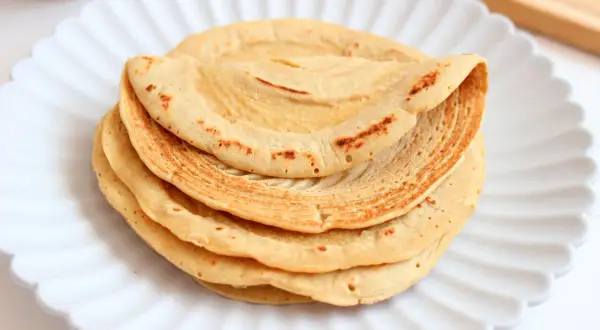 Pila di piadine di ceci dorate e leggermente maculate, servite su un piatto bianco a forma di fiore.
