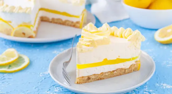 Una fetta di torta fredda al limone, con strati cremosi e una gelatina gialla brillante, decorata con ciuffi di panna e fette di limone.