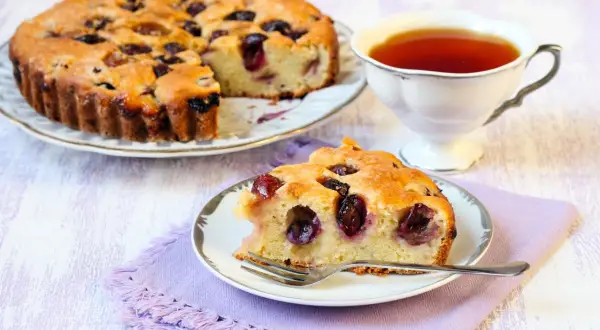Una fetta di torta bertolina, soffice e profumata, con ciliegie mature, accanto a una tazza di tè fumante.