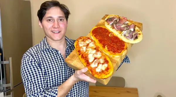 Uomo sorridente mostra tre lingue di pizza senza lievito: una margherita, una al pomodoro e una con prosciutto e funghi.