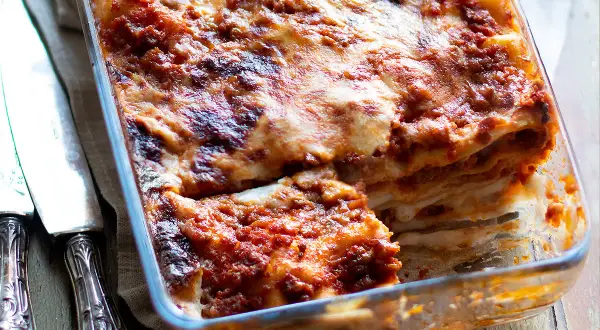 Lasagna al ragù appena sfornata, dorata e filante, in una teglia di vetro, con posate d'argento a lato.