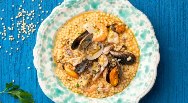 Piatto di fregola con pesce, cozze, vongole e gamberi, guarnito con prezzemolo tritato.