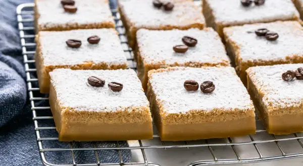Fette di torta magica al caffè, spolverate di zucchero a velo e decorate con chicchi di caffè, riposano su una griglia.