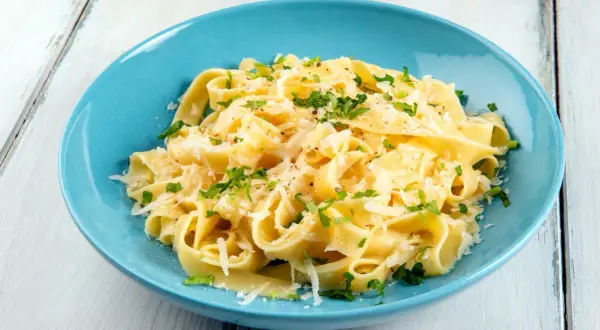 Un piatto di pasta burro e parmigiano, condita con prezzemolo fresco, servita in una ciotola blu su un tavolo di legno.