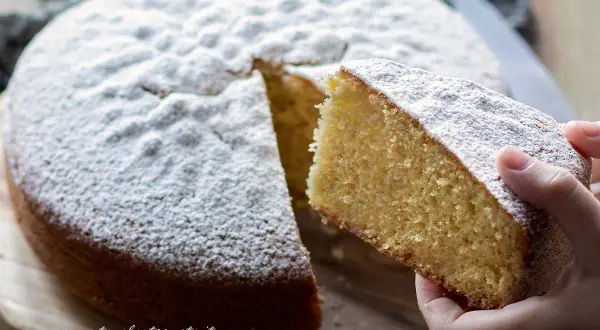 Una fetta di torta margherita soffice e profumata, cosparsa di zucchero a velo, viene sollevata da una mano.
