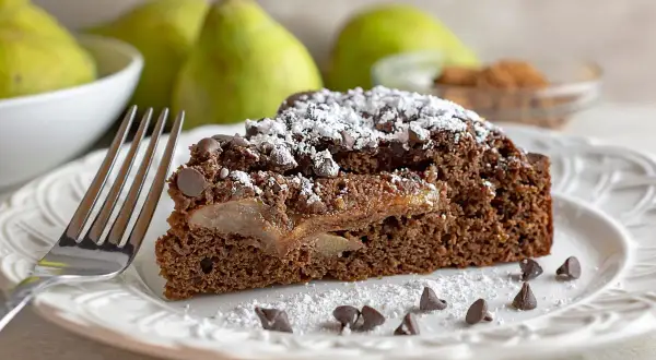 Una fetta di torta pere e cioccolato, cosparsa di zucchero a velo e gocce di cioccolato, con pere caramellate all'interno.