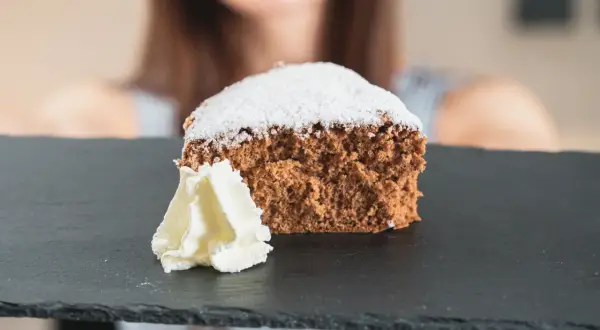 Una fetta di pan di spagna al cacao, soffice e spolverata di zucchero a velo, servita con panna montata su una lavagna scura.