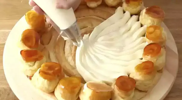 Una mano decora una torta con crema chiboust, creando un motivo a ventaglio. Piccoli bignè caramellati circondano la base.