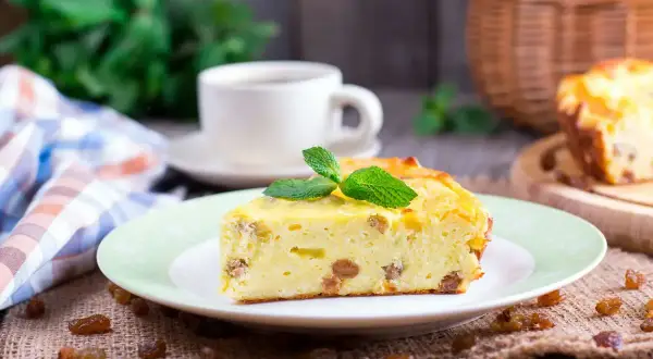 Una fetta di torta putana, soffice e dorata, con uvetta e una fogliolina di menta, accanto a una tazza di caffè.