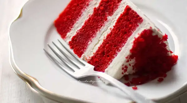 Una fetta di torta red velvet, stratificata con crema bianca, su un piatto bianco con una forchetta.
