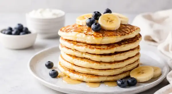 Pila di pancake proteici guarniti con mirtilli e banane, inzuppati nello sciroppo.