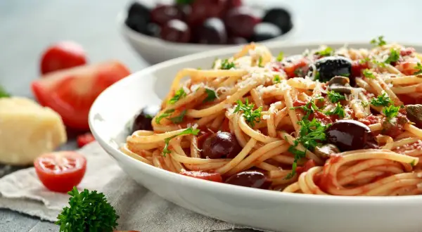 Un piatto di spaghetti alla puttanesca, conditi con pomodoro, olive e capperi, guarnito con prezzemolo e parmigiano.