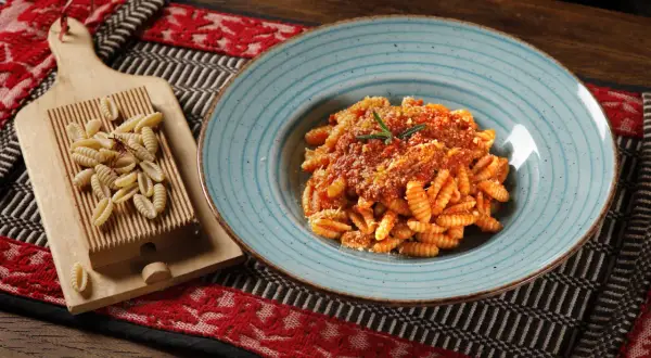 Gnocchetti sardi conditi con sugo rosso e parmigiano, accanto a una spianatoia con pasta fresca.
