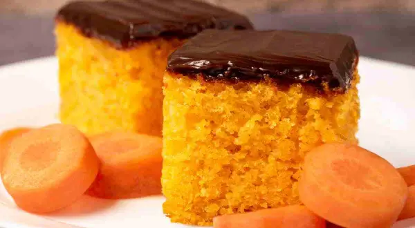 Due fette di torta di carote, soffice e arancione, ricoperte da glassa al cioccolato fondente. Accanto, fette di carota fresca.