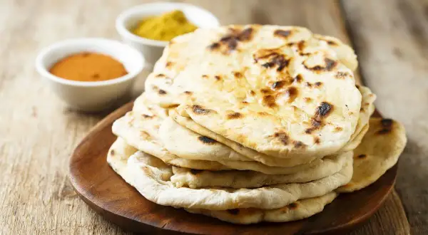 Pila di pane naan appena sfornato, con due ciotoline di spezie in sottofondo.