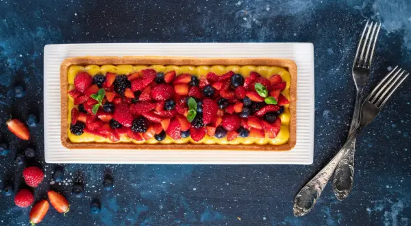 Una crostata di frutta moderna, con crema gialla e un tripudio di fragole, mirtilli e more fresche, guarnita con menta.
