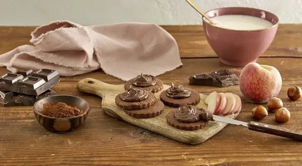 Biscotti senza burro al cacao, guarniti con crema al cioccolato, su tagliere di legno. Ingredienti: cioccolato, nocciole, pesca, latte.