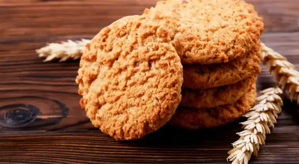 Biscotti integrali dorati e croccanti, impilati su un tavolo di legno scuro, con spighe di grano a decorare.