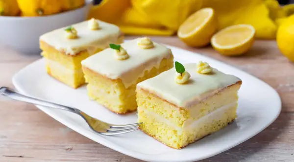 Tre fette di torta al limone e cioccolato bianco, guarnite con glassa e foglioline verdi, su un piatto bianco con limoni sullo sfondo.