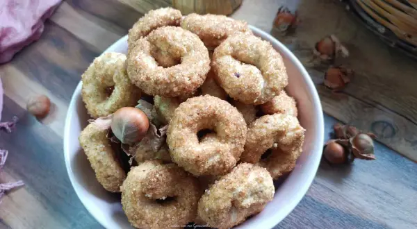 Ciambelline al vino cosparse di zucchero, con nocciole e un drappo rosa sullo sfondo.