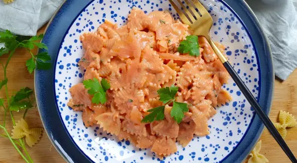Farfalle al salmone cremose, guarnite con prezzemolo fresco, servite su un piatto blu e bianco con una forchetta dorata.