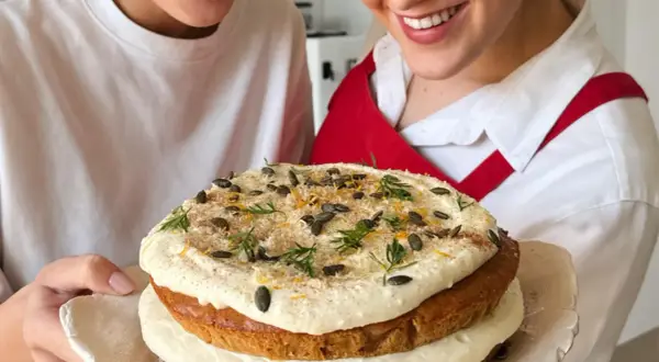 Due persone sorridono mostrando una torta a strati decorata con glassa, semi di zucca e scorza d'arancia. La ricetta della carrot cake è un successo!