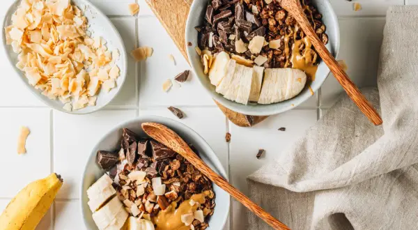 Due ciotole con granola fatta in casa, banana a fette, cioccolato fondente e burro d'arachidi. A lato, scaglie di cocco tostato.