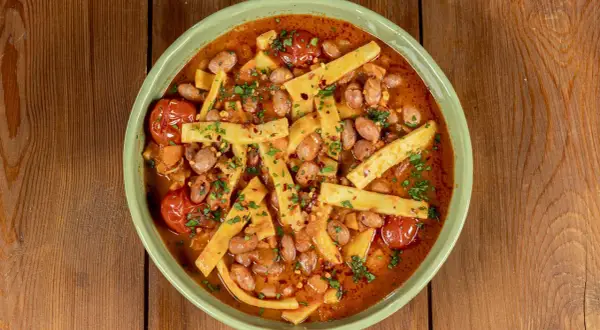 Piatto di sagne e fagioli fumante, con pomodorini e prezzemolo tritato, servito in una ciotola verde su un tavolo di legno.