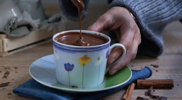 Una mano versa cioccolata calda in una tazza decorata con fiori. Cannella e scaglie di cioccolato completano la scena.
