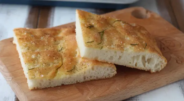 Focaccia senza glutine dorata e fragrante, condita con sale grosso e rosmarino, su un tagliere di legno.