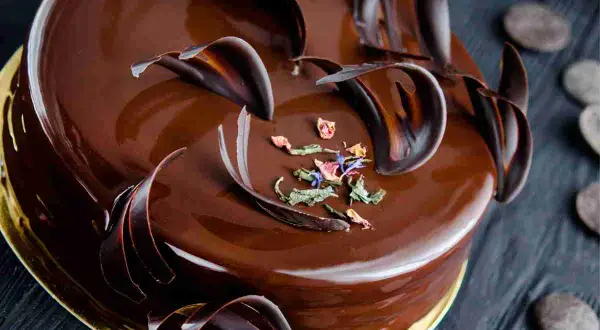 Una lucida torta Savoia al cioccolato, decorata con riccioli di cioccolato fondente e petali di fiori secchi.
