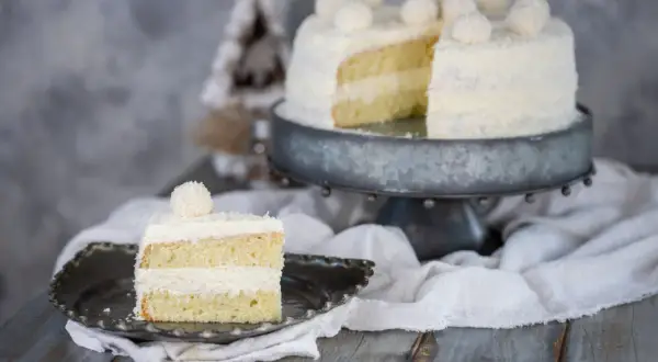 Una fetta di torta Raffaello, soffice e ricoperta di cocco, su un piatto decorato. Sullo sfondo, la torta intera su un'alzata.