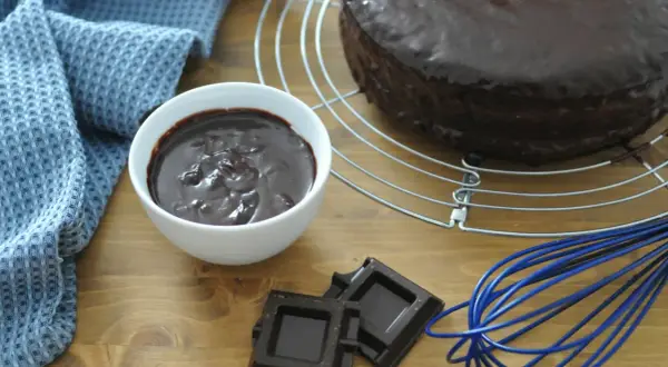 Torta ricoperta di lucida ganache cioccolato per copertura, con ciotolina di ganache, cioccolato fondente e frusta blu.