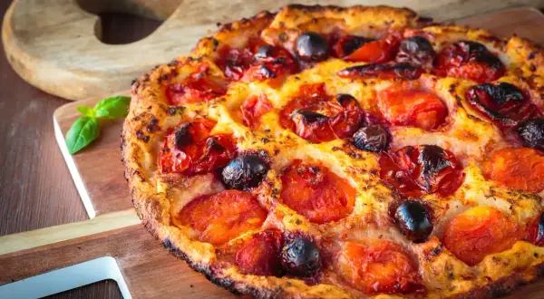 Focaccia pugliese dorata con pomodorini succosi e olive nere, guarnita con basilico fresco su tagliere di legno.