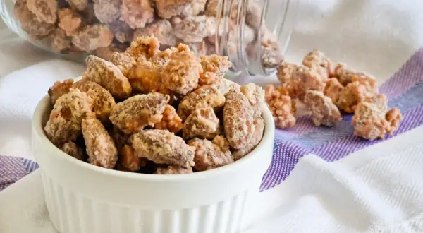 Un bianco contenitore pieno di mandorle pralinate, con altre sparse su un tovagliolo a righe.