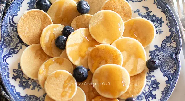 Mini pancake ricetta: tanti piccoli pancake dorati, guarniti con mirtilli freschi e sciroppo d'acero, serviti su un piatto decorato.