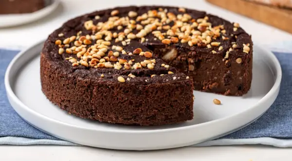 Torta paesana al cioccolato, ricca e umida, guarnita con pinoli tostati. Una fetta è stata già servita, invitando a gustare questo dolce rustico.