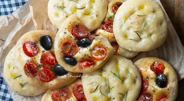 Focaccine in padella, alcune condite con pomodorini e olive nere, altre con rosmarino e olio.
