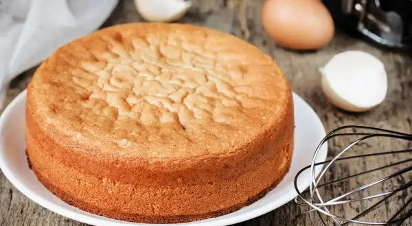 Torta 12 cucchiai soffice e dorata, pronta per essere decorata. Ingredienti per la torta 12 cucchiai sparsi sul tavolo: uova, frusta.