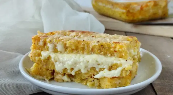 Una fetta di pasticcio di polenta con formaggio filante, cotto al forno e servito su un piatto bianco.