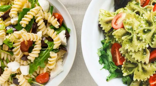 Due insalate di pasta fredda senza pomodori: una con fusilli, melanzane e mozzarella, l'altra con farfalle al pesto e pomodorini.