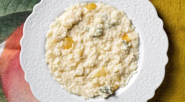 Un cremoso risotto pere e gorgonzola, servito su un piatto bianco decorato, con pezzetti di pera e formaggio blu visibili.
