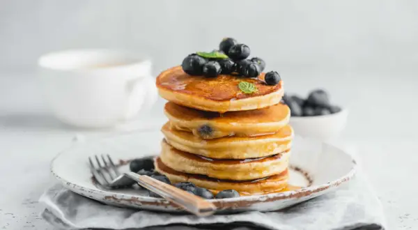 Pila di pancake con farina di riso, guarniti con mirtilli freschi e sciroppo d'acero, su un piatto rustico con una tazza di caffè sullo sfondo.