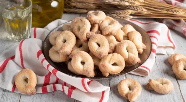 Un piatto colmo di taralli al vino bianco, cosparsi di zucchero, accanto a un bicchiere di vino e una bottiglia d'olio.