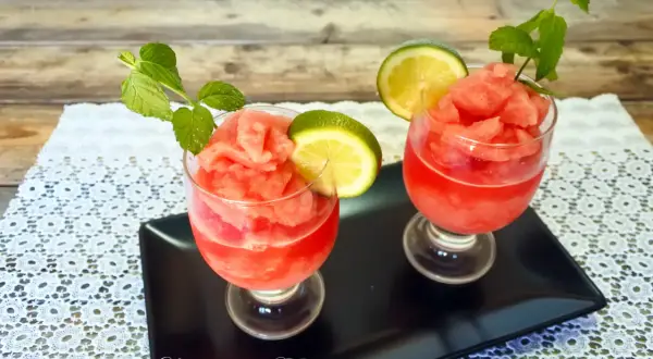 Due bicchieri di granita di anguria, guarniti con fettine di lime e foglie di menta, su un vassoio nero.