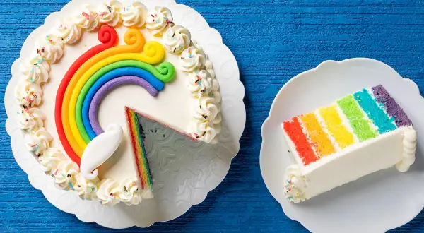 Torta arcobaleno con strati colorati e decorazioni di panna. Una fetta è già servita su un piattino.