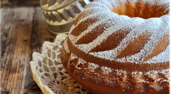 Ciambellone soffice come una nuvola, cosparso di zucchero a velo, adagiato su un centrino di pizzo, su un tavolo di legno rustico.