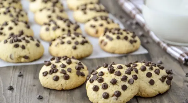 Biscotti con gocce di cioccolato, dorati e invitanti, sparsi su un tavolo di legno. Alcuni sono in primo piano, altri sullo sfondo.